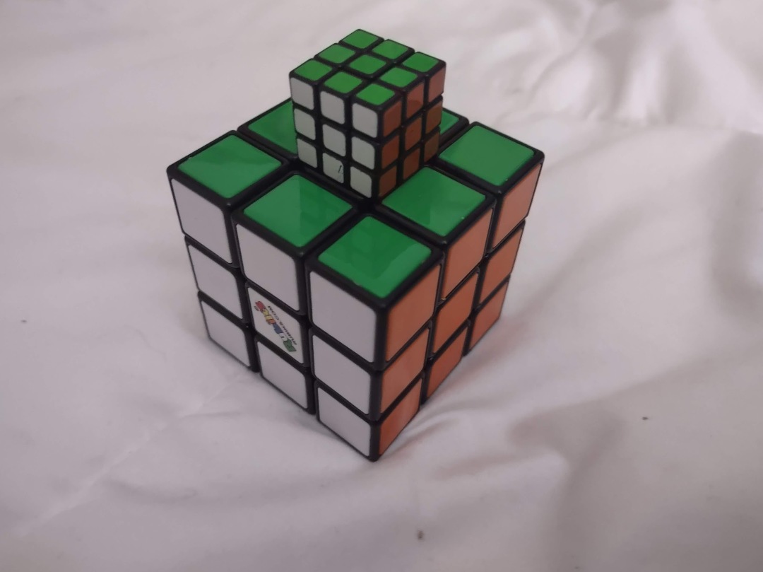 a very small rubik's cube on top of a larger rubik's cube. The smaller cube is the size of one tile of the larger cube.