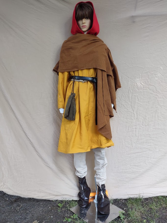 A mannequin wearing a yellow tunic over tan hose and dark brown boots as well as a belt, pouch, and red hood and also a dark tan short cloak.