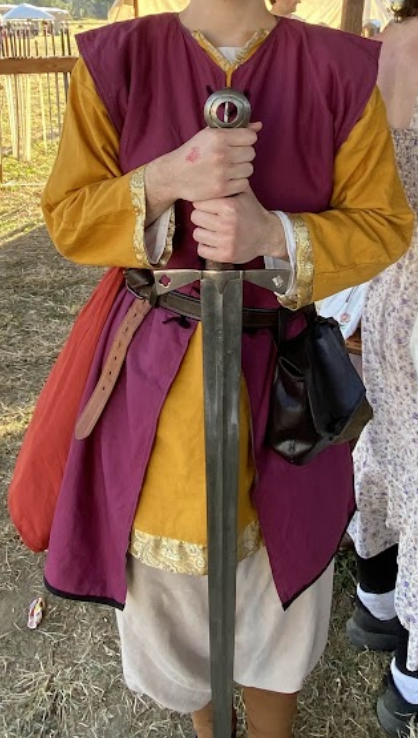 a picture of a medieval-style outfit. There is a trimmed yellow tunic, a maroon overtunic, a leather belt with a belt pouch, and loose pants with wrapped lower legs. The person wearing the outfit has their head cropped out and is holding a sword.